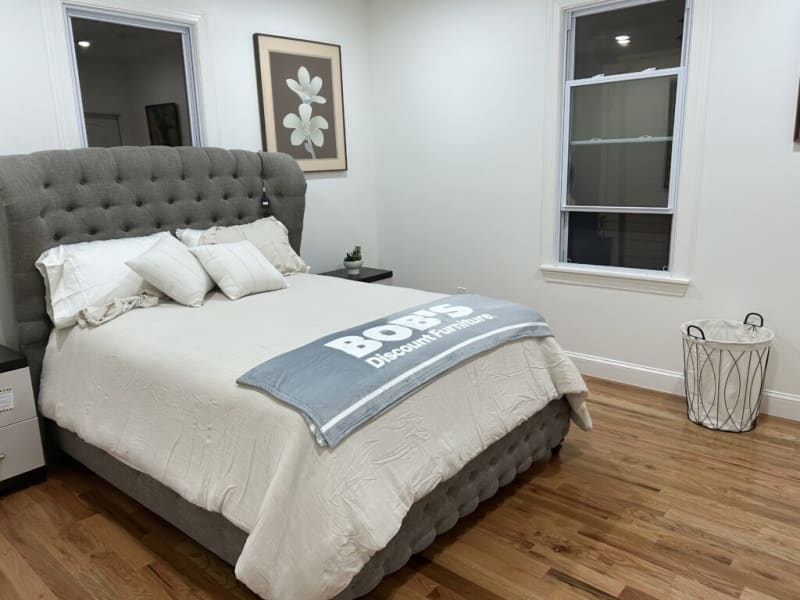 Queen-sized tufted gray upholstered bed with beige bedding, decorative pillows, and a gray throw blanket reading "BOSS Discount Furniture" in a minimalist bedroom with hardwood floors.