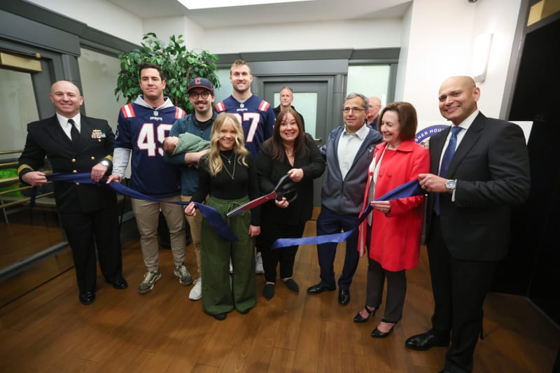 Group ribbon-cutting ceremony with diverse participants including a woman holding large scissors, people in business attire, and two men in New England Patriots jerseys.