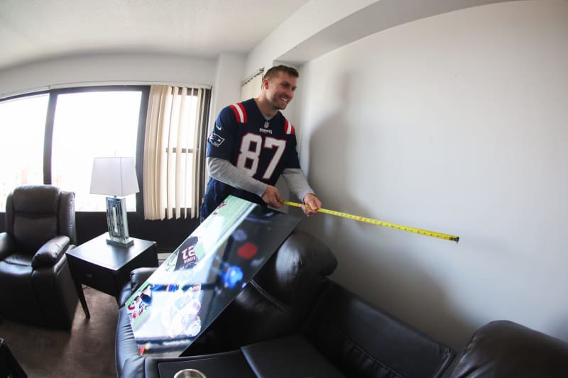 Man measuring wall space with a yellow tape measure in a living room, holding a framed sports poster featuring a football player wearing a New England Patriots jersey number 87.