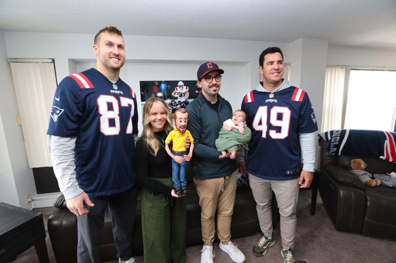 Three adults and two children stand in a living room with sports-themed decor. Two adults wear navy blue New England Patriots jerseys (#87 and #49) with white long sleeves underneath. One adult wears a dark jacket and tan pants, holding a baby wrapped in a green blanket. A woman in black top and green pants holds a child wearing a yellow shirt and blue jeans. A New England Patriots blanket and teddy bear are visible on a leather chair.