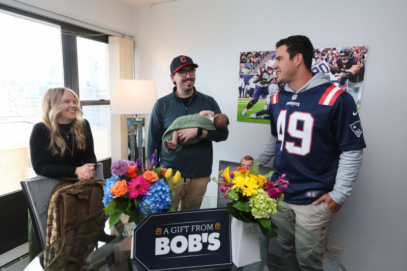 Three adults stand around a glass table with two vibrant floral arrangements in blue, orange, yellow, and pink hues. One person holds a baby wrapped in a green blanket. A sign on the table reads "A GIFT FROM BOB'S." The setting is a bright room with large windows and a sports-themed wall art featuring a football game. One adult wears a navy Patriots jersey with number 49.