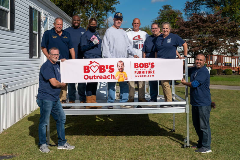 Group of people holding a banner that reads "BOB'S Outreach | BOB'S Discount Furniture MYBOBS.COM" outdoors on a sunny day.