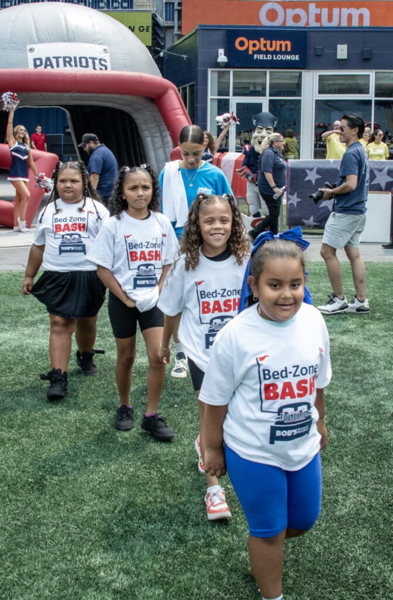 Children wearing white "Bed-Zone BASH" t-shirts and athletic shorts line up on artificial turf at a Patriots-themed event near the Optum Field Lounge.