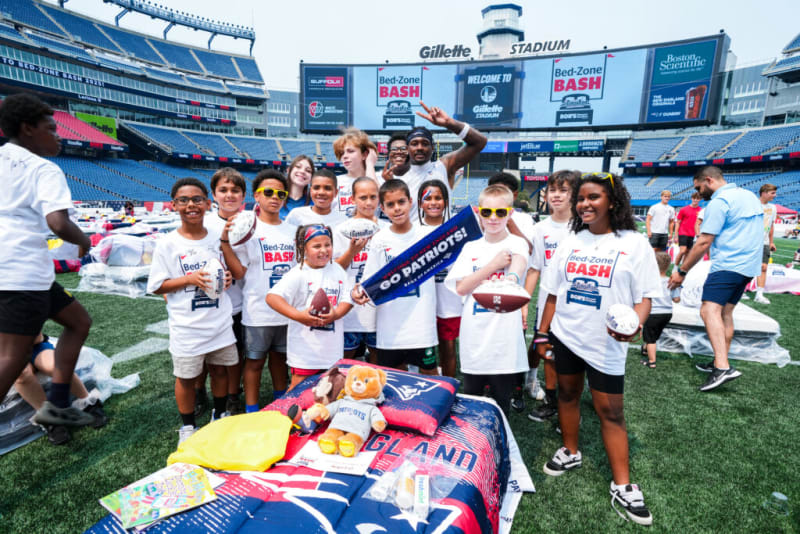 Group of children and adults wearing white Bed-Zone Bash T-shirts holding footballs and a "Go Patriots!" banner on the field at Gillette Stadium.