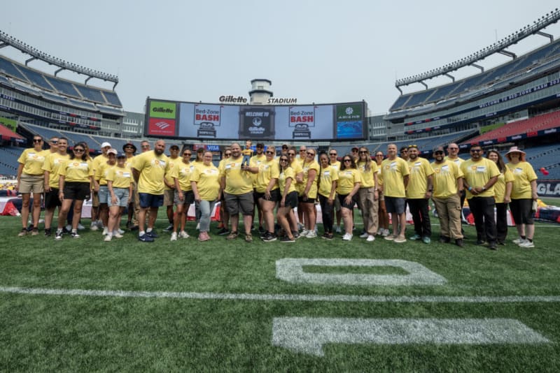 Group of people wearing matching yellow t-shirts standing on the field at Gillette Stadium with large screens displaying "Bed-Zone Bash" event branding.