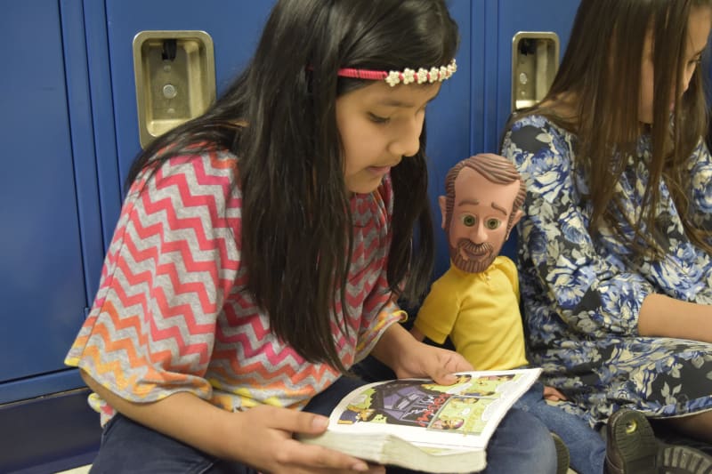 Two children sitting by blue lockers, one wearing a colorful zigzag patterned shirt reading a comic book with visible cartoon panels, the other in a yellow shirt and jeans.