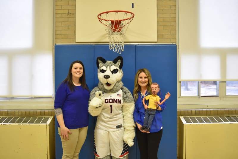 Two women stand in a gymnasium next to a UConn husky mascot wearing a white jersey with "UCONN" and number 1, under a basketball hoop. One woman holds a puppet.