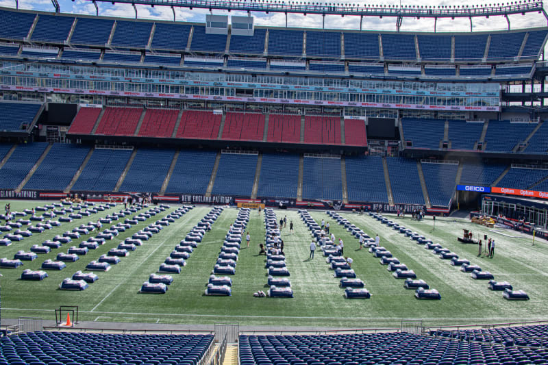 A large football stadium with rows of medical beds spaced evenly on the field, labeled with New England Patriots branding, likely set up for a mass vaccination or medical event.