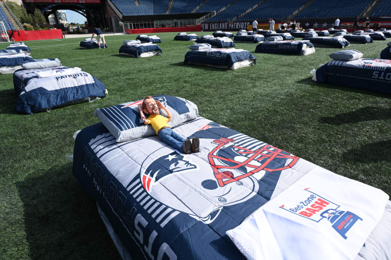 Single bed with New England Patriots-themed bedding and pillow on a football field, featuring a "BedZone Bash" towel for a fan event.