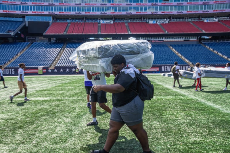 A person carrying large plastic-wrapped bundles on their head walks on a football field with New England Patriots stadium seats in the background.
