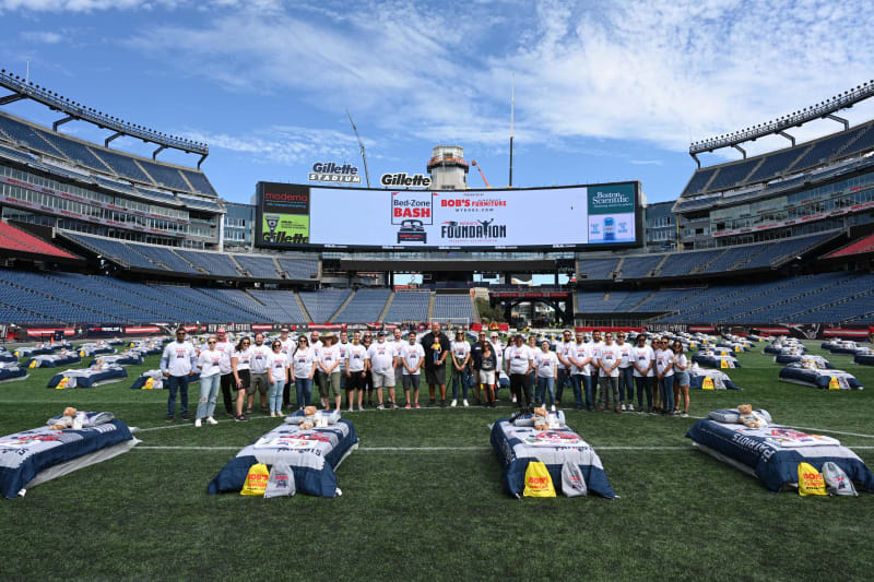 Group of people standing on a football field at Gillette Stadium with multiple beds set up, featuring BOB'S Furniture and Bed-Zone Bash branding on bedding and bags.