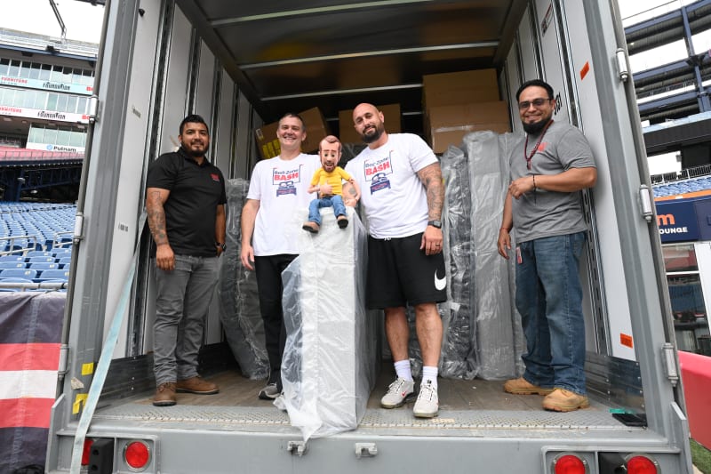 Five men standing inside the back of a moving truck filled with wrapped furniture and boxes. Two men wear white t-shirts with "Bed-Zone BASH" and a graphic of a bed on them, indicating a promotional or charity event. The setting appears to be a stadium or large outdoor venue.
