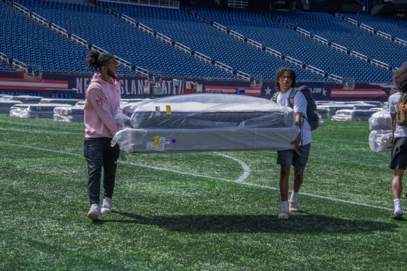 Two people carrying a wrapped mattress and box spring across a green sports field with empty blue stadium seats in the background.