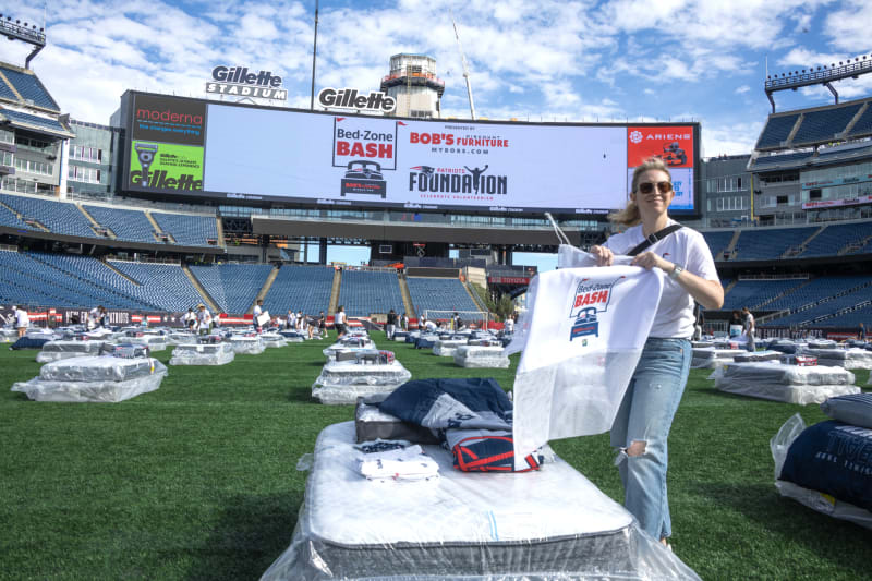 Gillette Stadium field set up with multiple beds covered in plastic, a person holding a white mesh bag with "Bed-Zone Bash" logo, and a large digital screen displaying "Bed-Zone Bash," "Bob's Furniture," and "Patriots Foundation Celebrate Volunteerism" text.