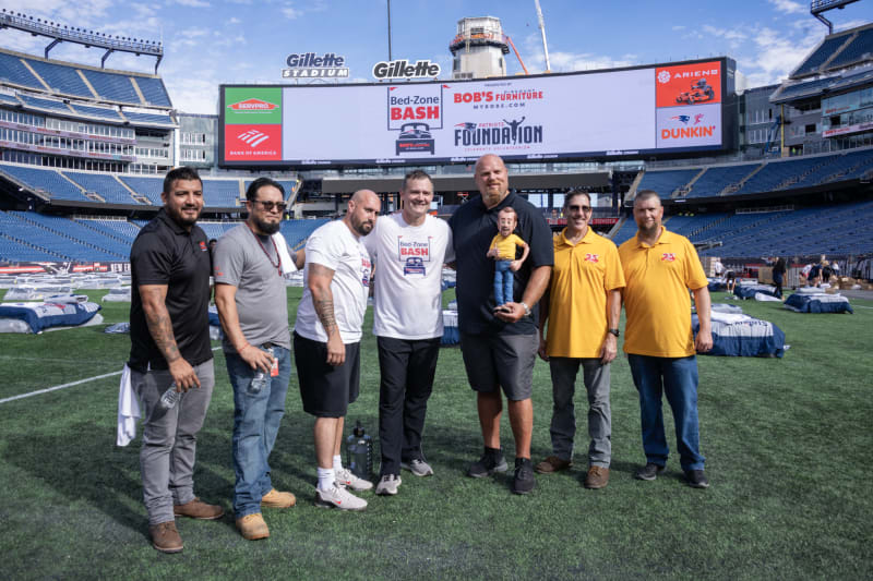 Seven men standing on a football field at Gillette Stadium, with a large digital scoreboard displaying "Bed-Zone Bash," "Bob's Discount Furniture," "Patriots Foundation," and sponsor logos including SERVPRO, Bank of America, Ariens, and Dunkin'. Two men wear yellow shirts with "25" logos, two wear white Bed-Zone Bash shirts, and others wear casual attire. Several beds are set up on the field in the background.