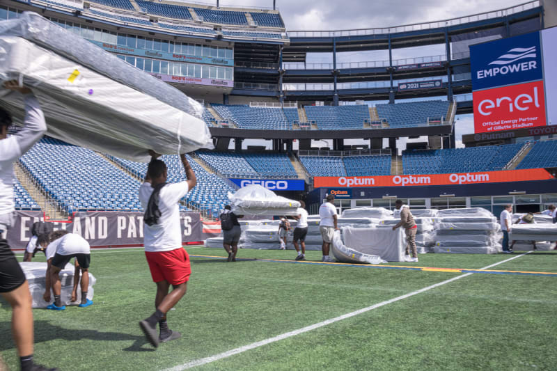 People carrying and organizing wrapped mattresses on the field of Gillette Stadium, with visible signage for Empower, Enel, Optum, and Geico sponsors.