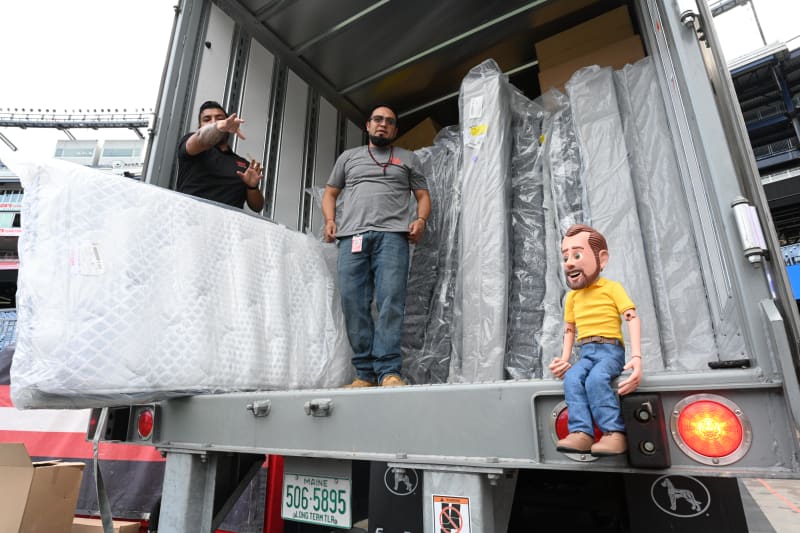 Two men unloading a truck filled with multiple wrapped mattresses and mattress toppers, one person sitting on the truck edge wearing a yellow shirt and jeans, Maine license plate 506-5895 visible.