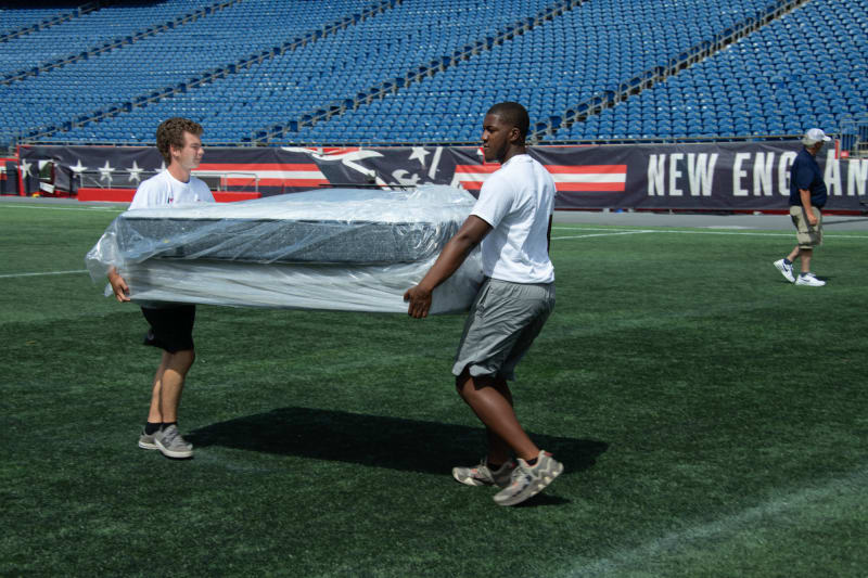 Two men in white shirts and shorts carry a large wrapped mattress on a green football field with blue stadium seats and a New England Patriots banner in the background.