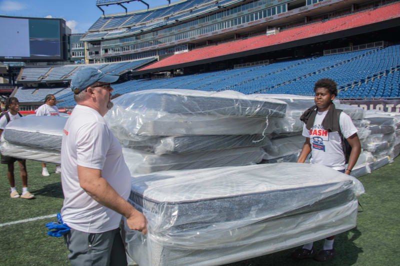 Two men carrying a wrapped mattress on a football field with stacks of plastic-covered mattresses and empty stadium seats in the background.