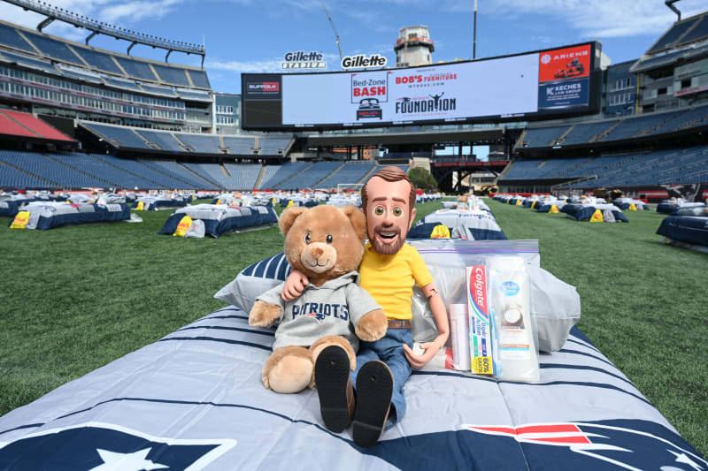 Stuffed teddy bear wearing a gray Patriots hoodie and a doll in a yellow shirt sitting on a striped Patriots bedspread at Gillette Stadium, with toiletries including Colgate toothpaste and shampoo in a clear plastic bag nearby.