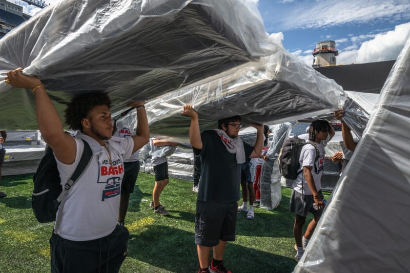 Young people wearing "Boys & Girls Club" t-shirts carry large, wrapped mattresses on a sports field under a blue sky, likely during a community event.
