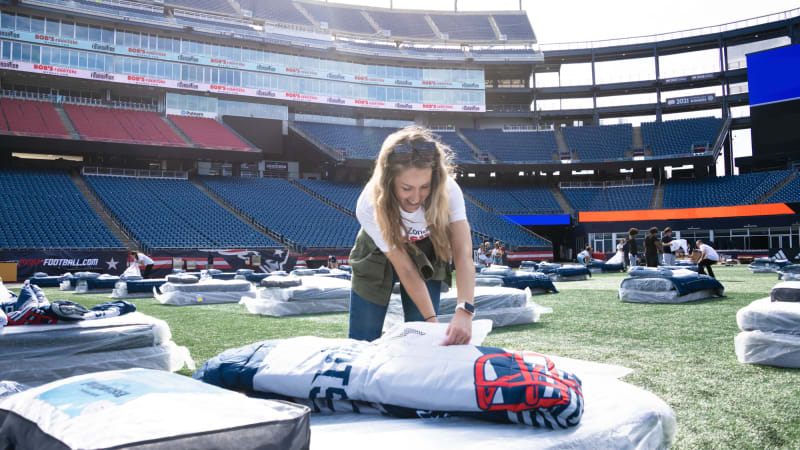 Person setting up a white and navy blue sleeping bag on artificial turf inside a large empty stadium with stadium seating and multiple sleeping bags scattered around.