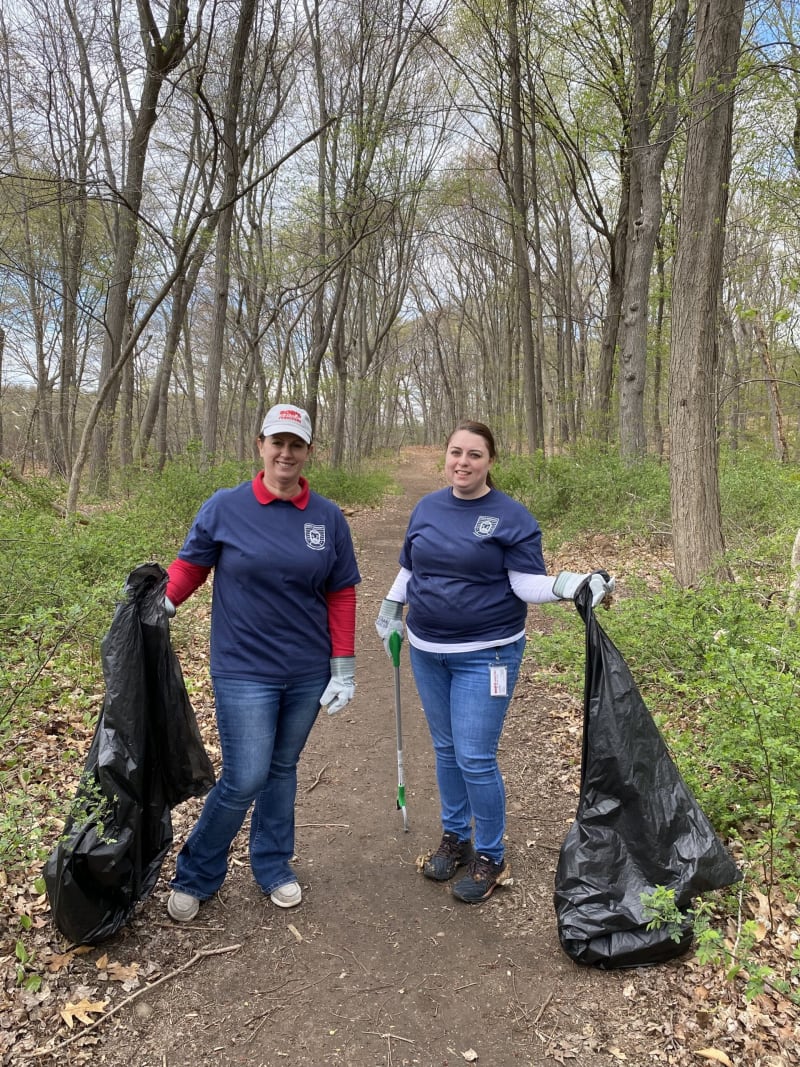 Two people wearing navy blue shirts and gloves holding large black trash bags and a litter picker, cleaning a wooded trail in early spring.