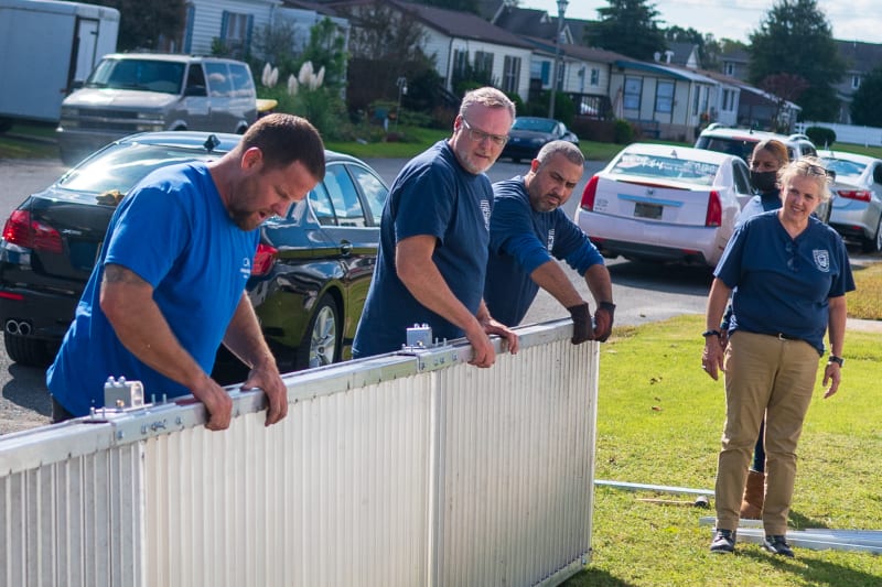 Group of people wearing blue shirts assembling or handling a white metal fence panel outdoors on a sunny day in a residential neighborhood.