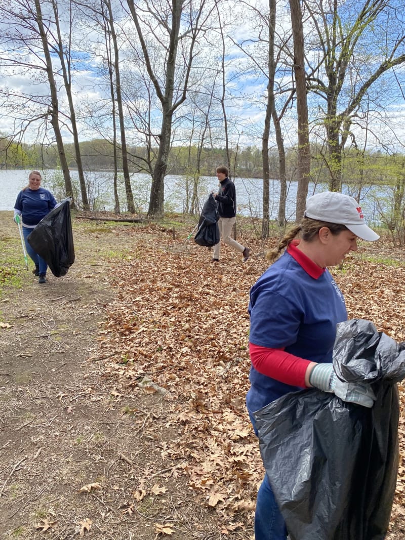 Volunteers wearing gloves and casual outdoor clothing collecting trash with black garbage bags in a wooded area near a lake on a clear day.