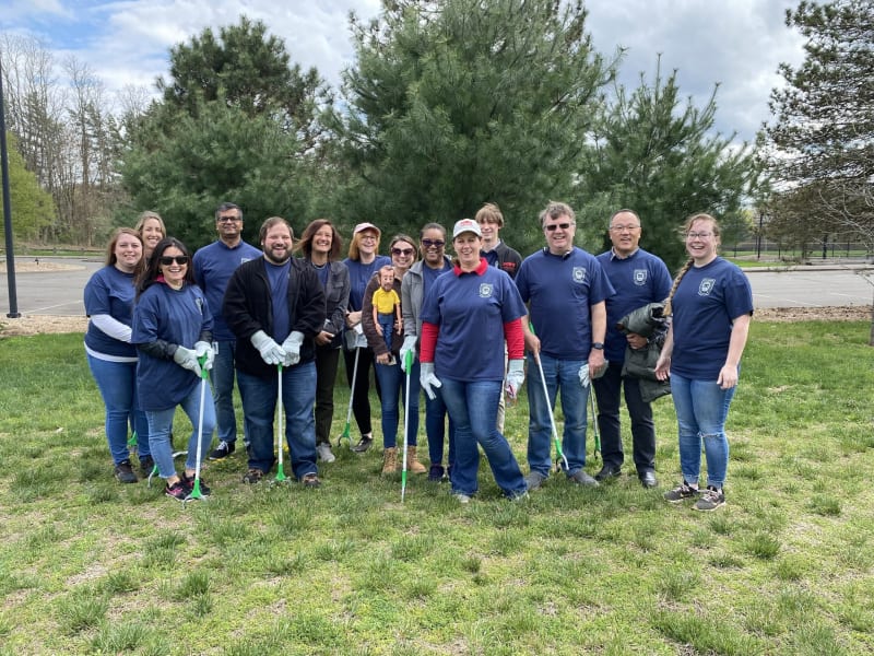 Group of people wearing navy blue shirts and jeans standing outdoors on grass with trees and a parking lot in the background, holding litter pickers and gloves for community cleanup.