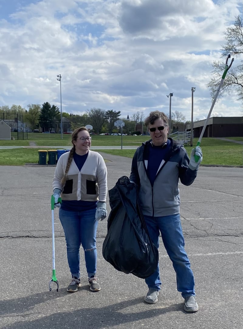 Two people wearing gloves holding trash grabbers and a large black garbage bag, participating in an outdoor cleanup on a paved area with grass and basketball hoops in the background.