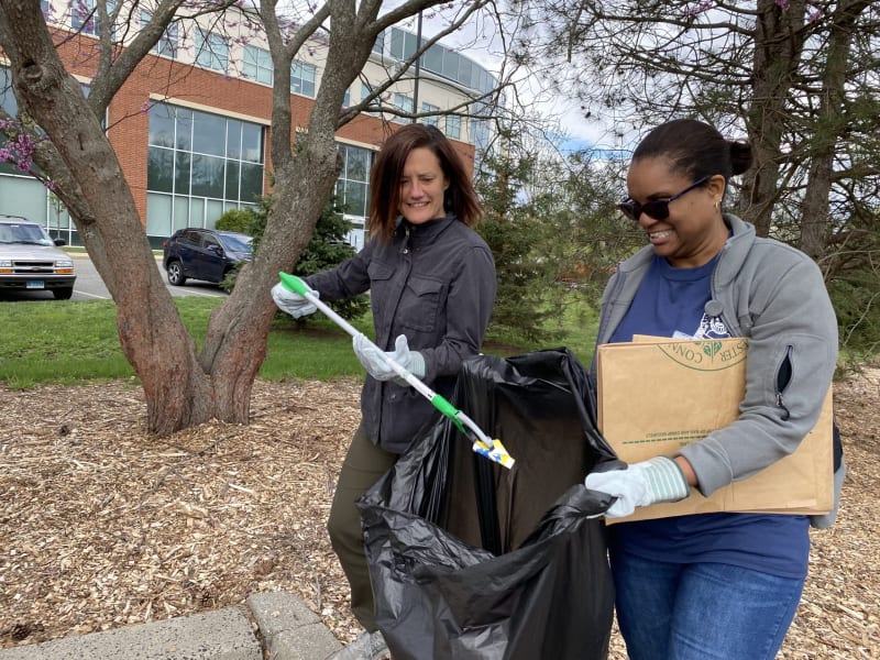 Two individuals wearing gloves use a grabber tool to pick up litter into a large black trash bag outdoors near trees and a building, promoting community cleanup and environmental care.