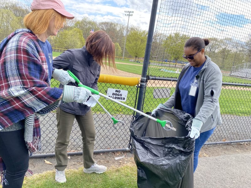 Three people wearing gloves use grabber tools to pick up trash into a large black garbage bag near a chain-link fence with a "NO DOGS ALLOWED" sign.