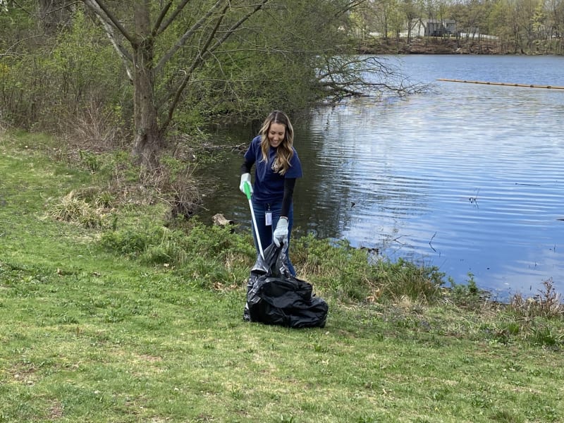 Person wearing gloves using a grabber tool to collect trash into a black garbage bag near a lake shore with green grass and trees.