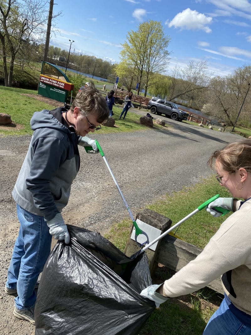 Two people wearing gloves use green-handled litter pickers to collect trash into a large black garbage bag at Union Pond Park on a sunny day with blue skies.