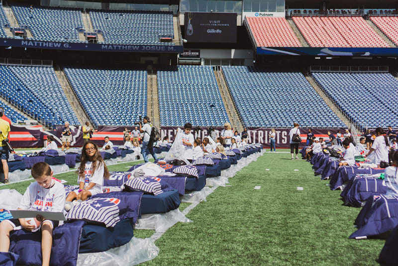 Rows of blue and purple cots with New England Patriots pillows set up on a football field, with children and adults interacting in a stadium with empty blue and red seats.