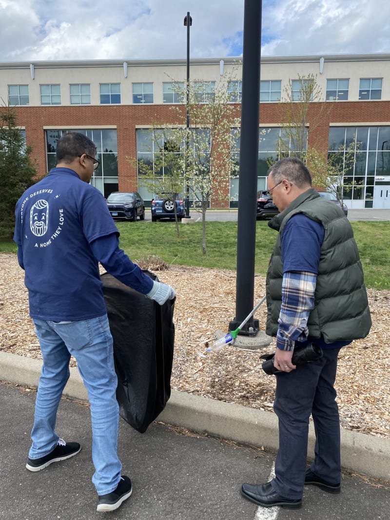 Two men wearing casual clothes and gloves are picking up litter outdoors; one holds a large black trash bag, the other uses a grabber tool to collect a plastic bottle.
