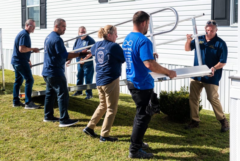 Group of six people carrying metal ramp sections outside a white building, wearing navy and blue shirts, assembling an accessibility ramp on grass.