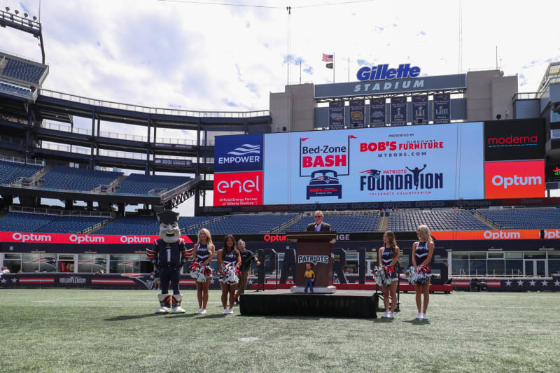 Gillette Stadium field setup with a podium, Patriots mascot, cheerleaders, and a large screen displaying sponsors including Empower, enel, Bob's Furniture, Patriots Foundation, Moderna, and Optum.