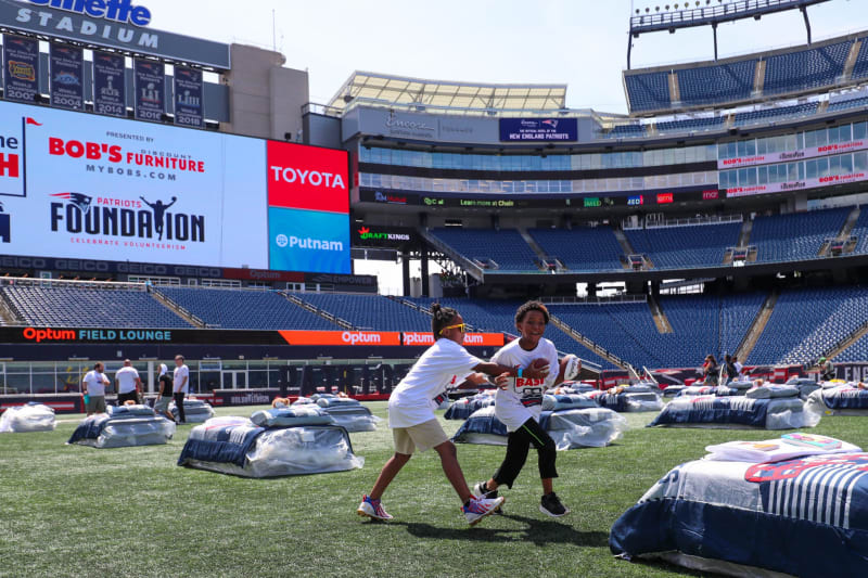 Two children playing football on the field at Gillette Stadium, with large sponsor screens displaying BOB'S FURNITURE, TOYOTA, and Putnam logos in the background.