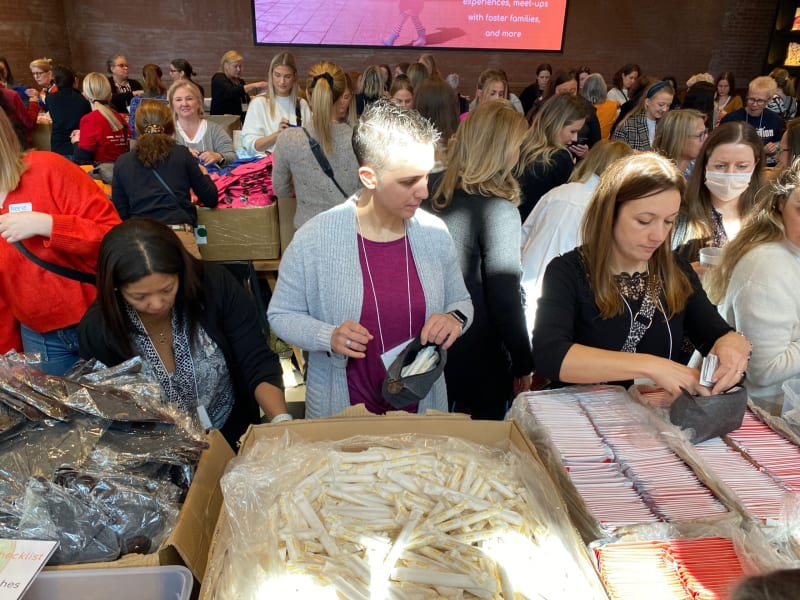 Volunteers at a busy event sorting and packing hygiene kits with individually wrapped items in cardboard boxes under bright lighting.