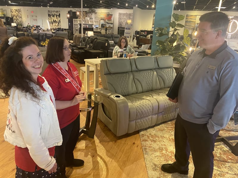Three people standing and conversing in a furniture store showroom with a gray leather reclining sofa featuring quilted stitching and cup holders in the center. The store has hardwood floors, various seating options, and modern decor.