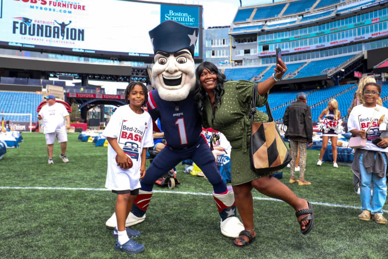 People posing on a football field with a person in a Patriots mascot costume. Two children wear white Bed-Zone Bash t-shirts, and a woman in a green dress takes a selfie. Stadium seats and cheerleaders are visible in the background.
