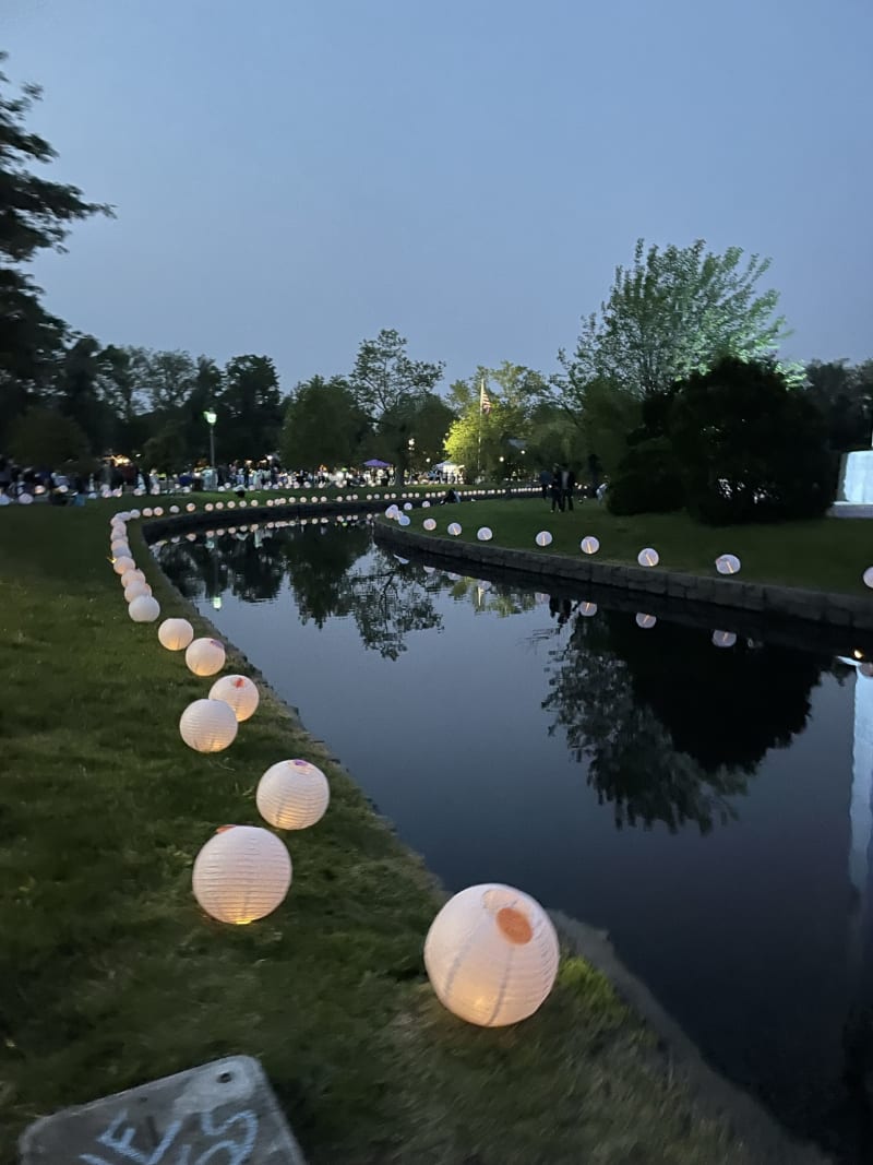 A winding canal bordered by glowing white paper lanterns on grassy banks at dusk, with trees and a crowd in the background.