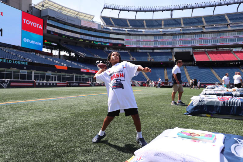 Child wearing a white "Bed-Zone Bash" t-shirt and black shorts throwing a football on a green turf field inside a large stadium with empty blue and red seats.
