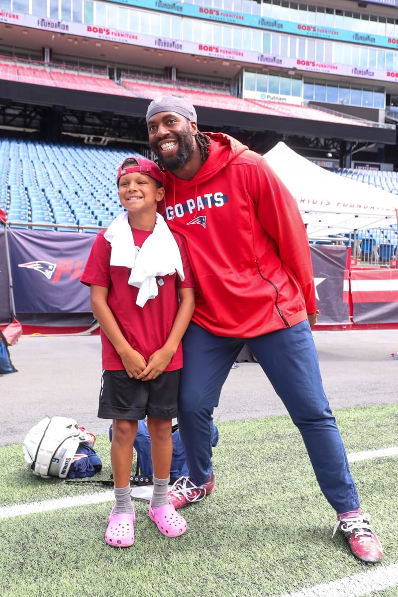 Two people standing on a football field near stadium seats; one wears a red "GO PATS" hoodie with navy pants and red cleats, the other wears a red t-shirt, black shorts, gray socks, and pink Crocs with a white towel draped over their shoulders.