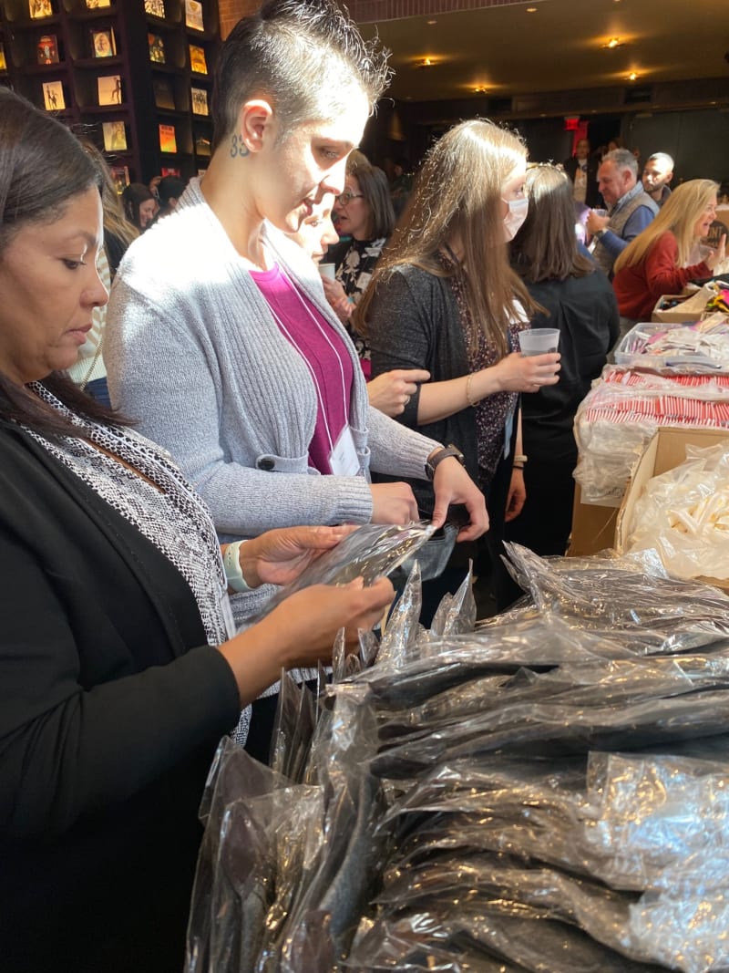 People browsing and selecting packaged clothing items at a busy indoor market or event, with stacks of plastic-wrapped apparel on the table.