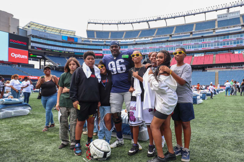 A group of diverse children and a football player wearing a navy jersey with number 96 pose together on a green field inside a large stadium with empty blue and red seats.