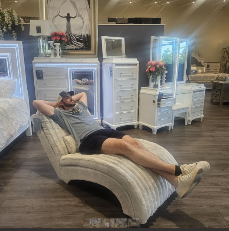 Man reclining on a plush, curved beige chaise lounge in a modern bedroom showroom with white wooden dressers, illuminated mirrors, and floral decor.