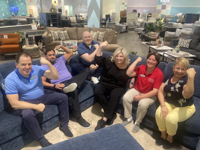 Group of six people sitting on blue fabric sectional sofas in a modern furniture store, flexing their arms in a playful, strong pose.